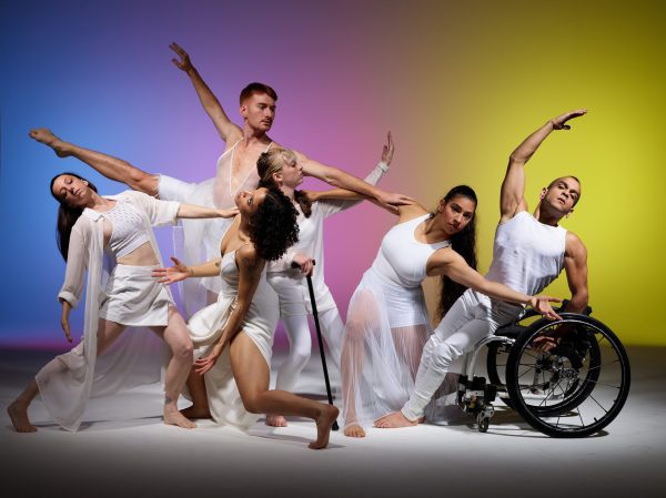 Six professional dancers from Axis Dance Company perform in flowing white costumes against a colorful gradient background. The dancers include both disabled and non-disabled performers - one dancer uses a wheelchair, another uses a crutch, while others are standing or in dynamic poses with arms extended. All are dressed in elegant white flowing garments, demonstrating the grace and artistry of inclusive contemporary dance.