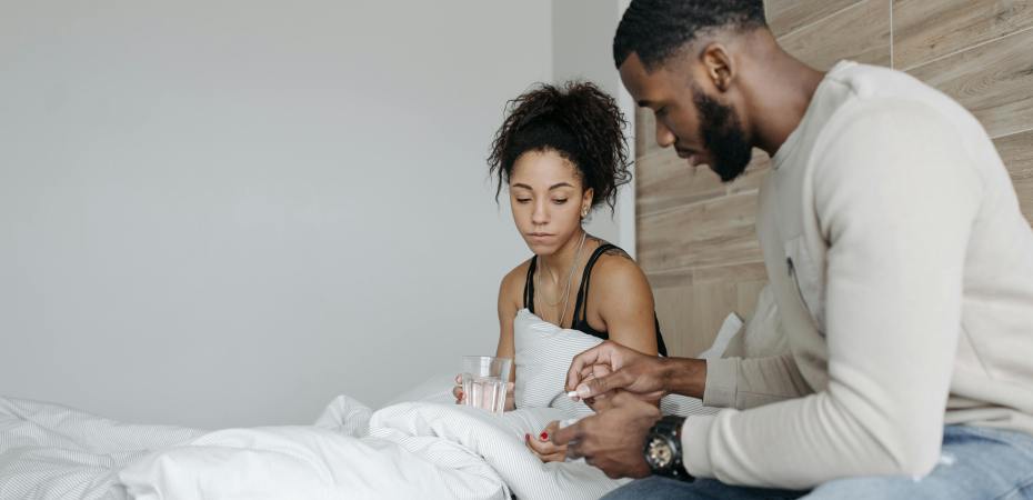 A man in a long-sleeved shirt is sitting on the bed beside a woman in a black tank top, and he's giving her medicine