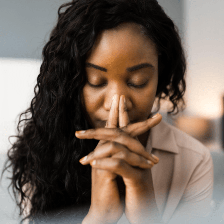 A close-up of a person with their eyes closed, hands clasped in front of their face, and dark curly hair.