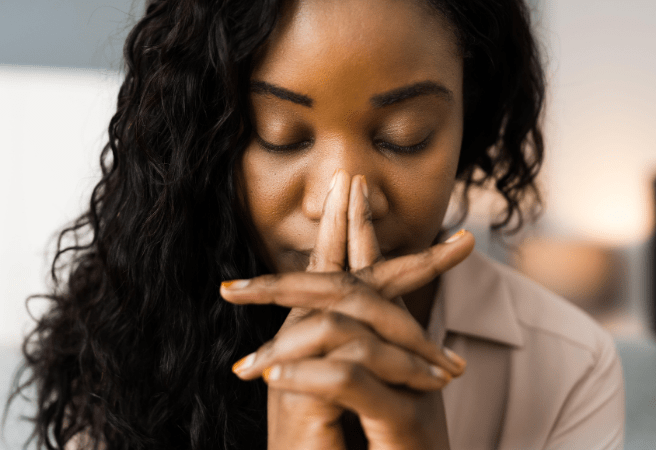 A close-up of a person with their eyes closed, hands clasped in front of their face, and dark curly hair.