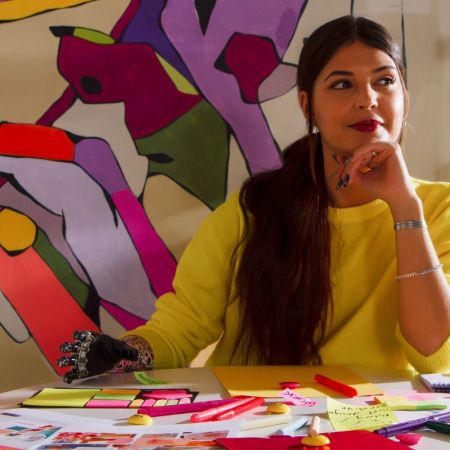 Woman with a prosthetic hand plots her next move, unbothered, in front of a chaotic and brilliantly colorful painting.
