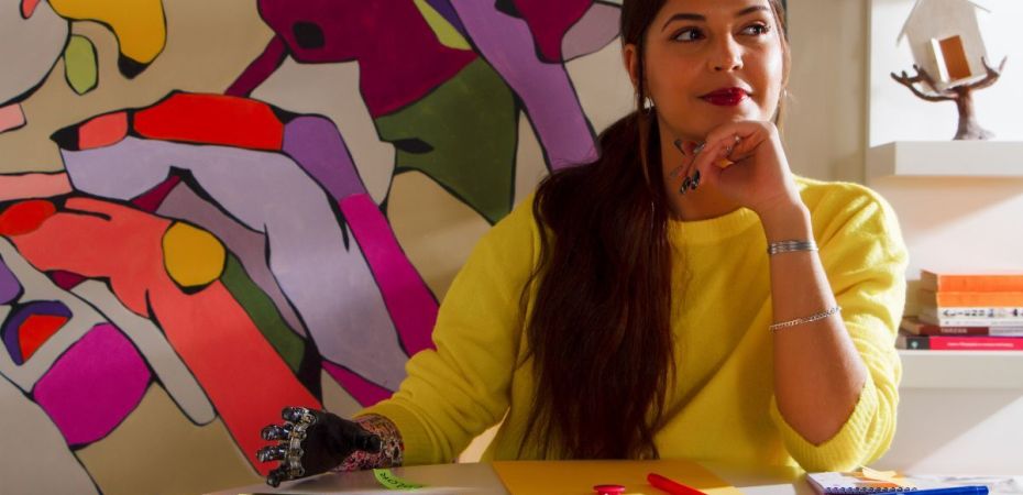 Woman with a prosthetic hand plots her next move, unbothered, in front of a chaotic and brilliantly colorful painting.