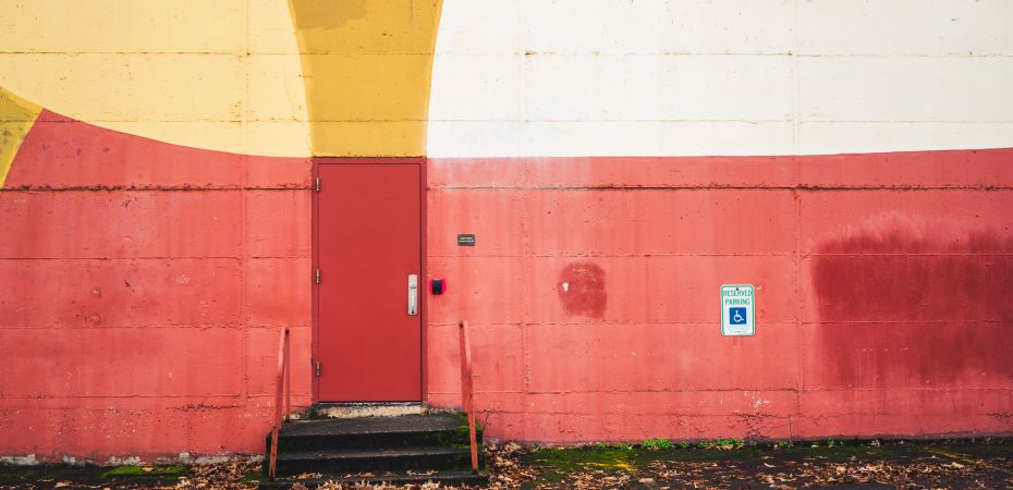 Accessible parking sign on a red wall beside a small entrance with steps leading up to a closed red door, with no ramp visible.